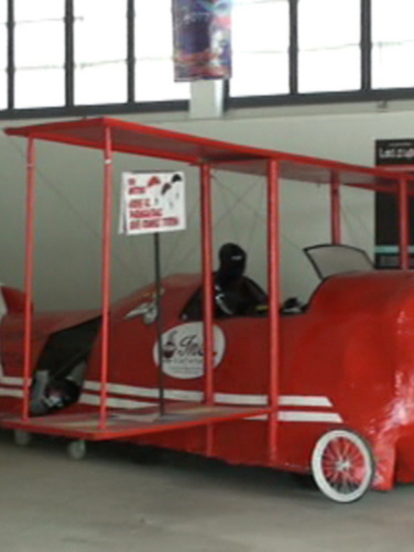 Un pequeño avión de juguete rojo con detalles blancos y azules está expuesto en un hangar. La imagen muestra una vista frontal del avión, con ruedas y asientos para dos personas. El fondo muestra una pared de ventanas y varios carteles colgados, lo que sugiere que este es un lugar dedicado a la aviación o una exposición de aviones.