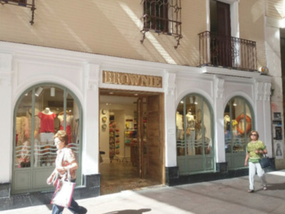 Tienda de ropa con maniquíes y escaparate visible. Edificio blanco con ventanas y balcones. Mujer caminando frente a la tienda.