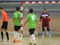 Un grupo de jugadores de fútbol en un campo interior, con dos jugadores en verde y uno en rojo. El portero está listo para recibir el balón, mientras que los otros jugadores esperan su turno. La imagen muestra un ambiente de juego en una cancha de futsal con una portería claramente visible.