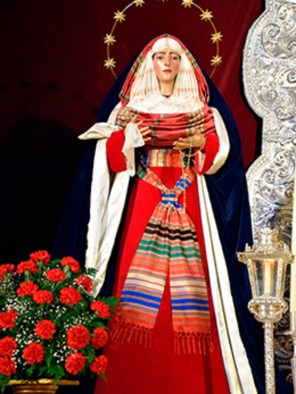 Una imagen de la Virgen María vestida con una túnica roja y un manto azul, rodeada de flores y adornos dorados.