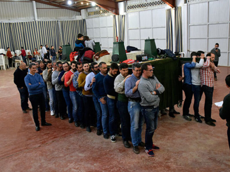 Una fila de hombres vestidos con camisas y pantalones, algunos con botas rojas, esperando en una sala amplia con paredes de azulejos y un suelo de baldosas.