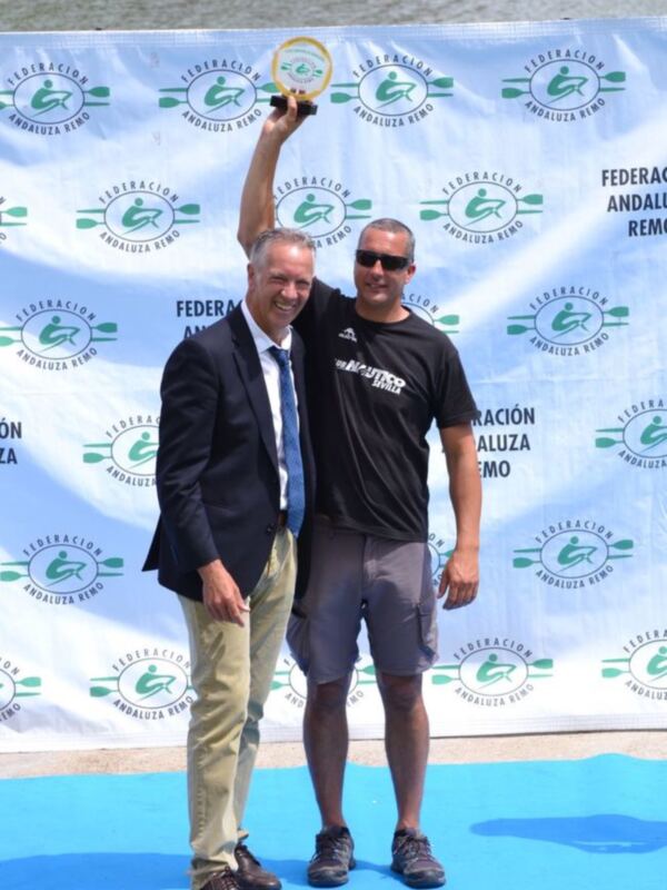 Dos hombres posan junto a un cartel de la Federación Andaluza de Remo, uno de ellos sostiene un premio.