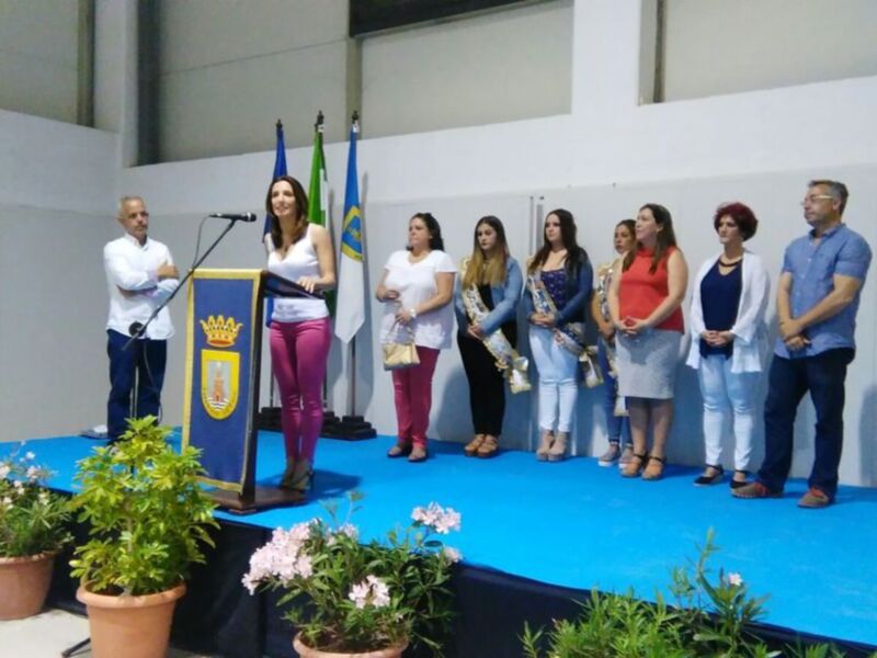 Una persona en el centro de la imagen está hablando frente a una audiencia. A su derecha, hay varios miembros de la audiencia, incluyendo una mujer con un bolso y otra con un vestido largo. En el fondo, se pueden ver banderas y una pantalla de proyección en la pared. La escena parece ser un evento formal o ceremonial, posiblemente una conferencia o presentación.