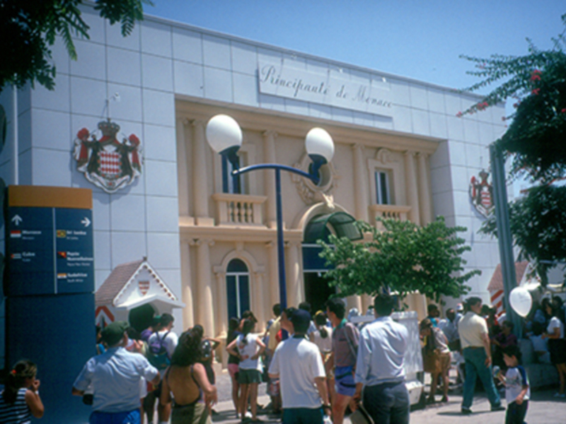 Gente caminando frente a un edificio blanco con un escudo y el texto 'Principauté de Monaco' en la fachada.