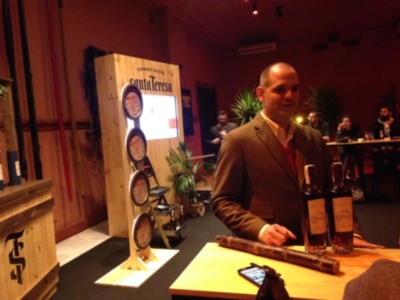 Un hombre en traje marrón sostiene tres botellas de vino en una mesa con un fondo de decoración y luces.