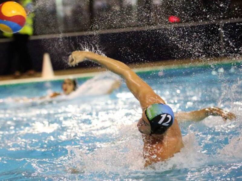 Un nadador en tronco de espalda, con una gorra de natación y un traje de baño blanco, se desplaza en una piscina azul. La persona está a punto de golpear el agua con su mano derecha, mientras que la izquierda se encuentra bajo el agua. La imagen captura un momento de intensidad en una competencia de natación, enfocándose en la técnica y el esfuerzo del nadador.