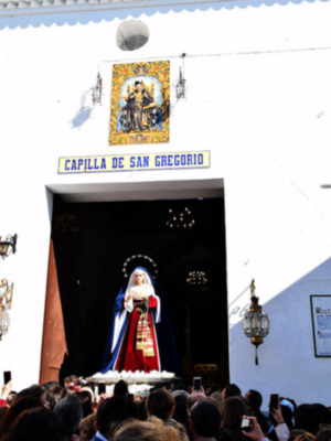 La Capilla de San Gregorio, con su arco y estandarte, alberga una imagen venerada de la Virgen María. Un grupo de personas observa con atención desde afuera, capturando el momento en sus teléfonos móviles.