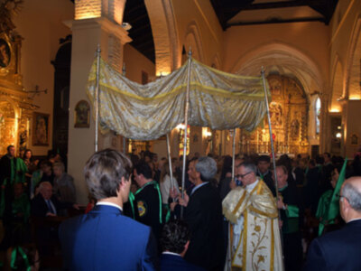 Una imagen de una iglesia llena de personas vestidas con trajes formales y capas coloridas, posiblemente durante una ceremonia religiosa o procesión. La arquitectura del edificio es clásica con columnas y bóvedas altas. La iluminación es dorada, reflejando la solemnidad del evento.