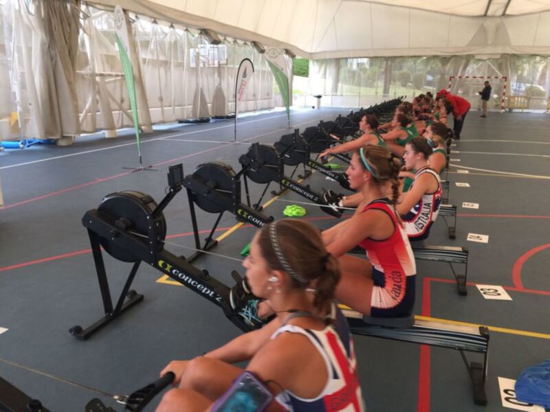 Una escena de entrenamiento indoor con mujeres en remo. Las personas están sentadas en remos Concept II, listas para un entrenamiento de remo. La imagen muestra una sala con múltiples remos y asientos, preparados para la práctica de remo. La iluminación es adecuada para un entorno indoor, y el ambiente parece ser una instalación de entrenamiento profesional.