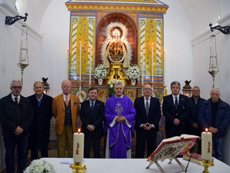 Se observa a una persona vestida con un hábito religioso, posiblemente un sacerdote, frente a una estatua de la Virgen María en el altar de una iglesia. A su lado, varios individuos vestidos formalmente observan la escena. La decoración del altar es colorida y elaborada, con flores blancas y detalles dorados. La iluminación es suave, destacando la figura central en el altar y los personajes de pie.