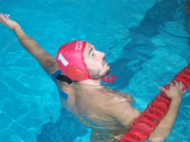 Persona nadando en una piscina, usando un gorro de natación rojo y brazalete, con el brazo extendido hacia arriba.