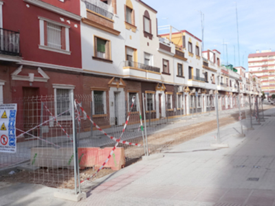 Una calle de viviendas en construcción o rehabilitación, con varios edificios en diferentes estadios de desarrollo. La calle está bloqueada por una cerca con señales de advertencia, indicando que hay trabajos en curso. La vivienda principal es roja con detalles blancos, mientras que las otras casas son de colores más suaves. La construcción se extiende a lo largo del camino, mostrando un proceso de urbanización en curso.