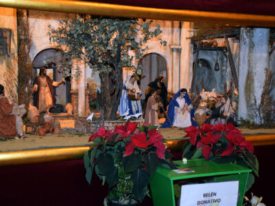 Belén de Navidad con escena de la Virgen y el Niño Jesús rodeados por los Reyes Magos. Detalle de una ofrenda en un contenedor verde con flores rojas y un letrero que dice "Belén Donativo".