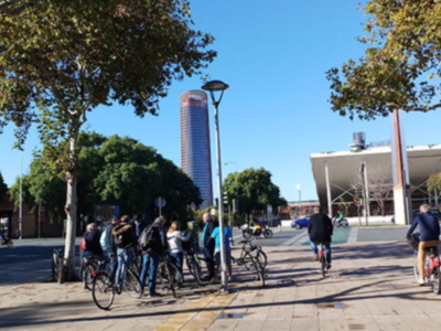 Una imagen de un parque con árboles, personas paseando y bicicletas. En el fondo, un edificio moderno y una torre de observación. El cielo es azul claro, indicando buen tiempo.
