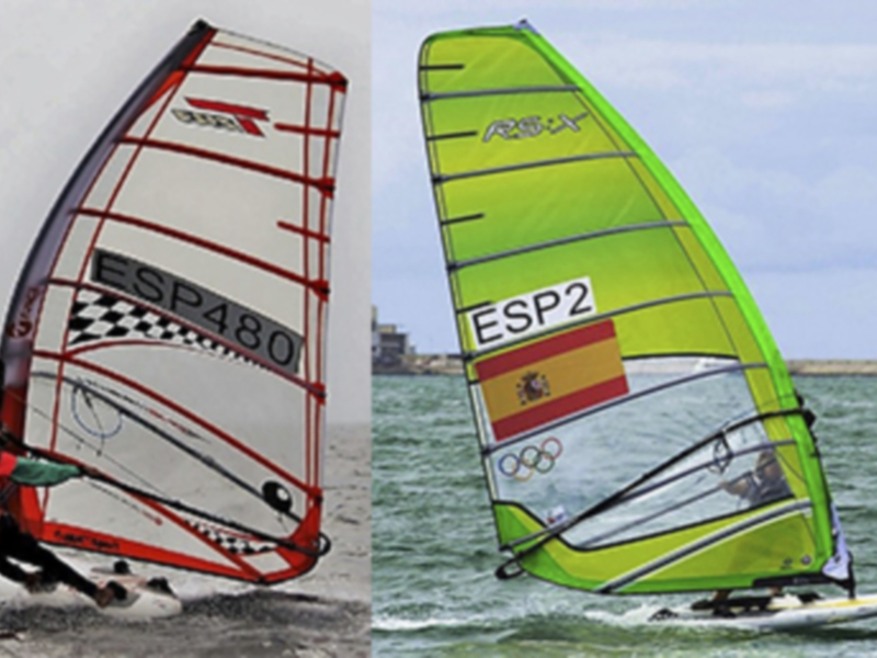 Dos personas practicando windsurfing en el mar, con una vela roja y otra verde. La vela roja tiene la inscripción "ESP480" y la vela verde tiene la bandera española con el símbolo olímpico. El fondo muestra un paisaje marino con una costa visible en el horizonte.