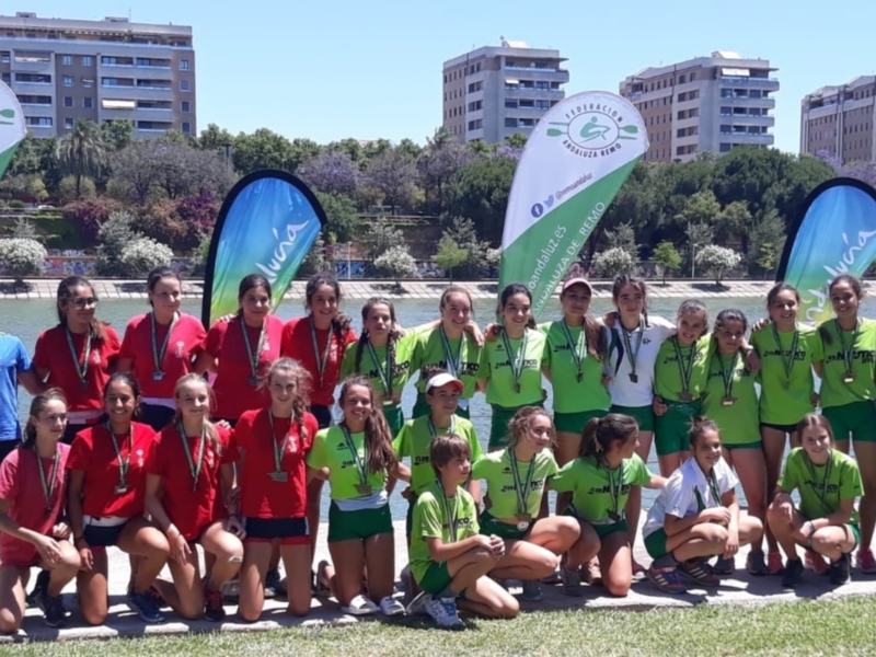 Equipo de atletas celebrando su victoria en el campeonato de Andalucía de Remo, con medallas en el cuello y banderas de la Federación Andaluza de Remo al fondo.