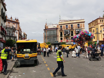 Un bus amarillo y negro está estacionado en un callejón de una ciudad con edificios históricos y gente caminando. El cielo está nublado, y hay un montón de globos de colores en el aire.