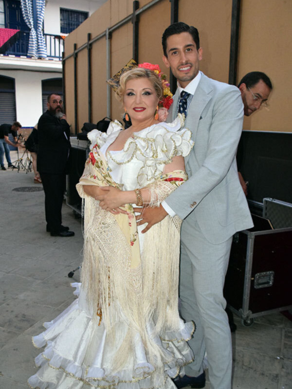 Un hombre y una mujer están posando juntos, con la mujer vestida con un traje de flamenca blanco y el hombre con una camisa azul y una corbata. La escena parece ser en un evento cultural o festivo, con otros asistentes en el fondo.