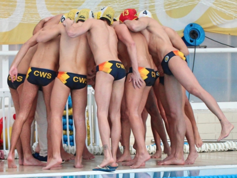 Una fila de nadadores en troncos de color negro y amarillo, con el logotipo "CWS" en la parte superior. Todos están listos para nadar, con sus piernas extendidas hacia adelante y pies apoyados en el borde de la piscina. El fondo muestra una barandilla blanca y un panel amarillo con letras blancas.