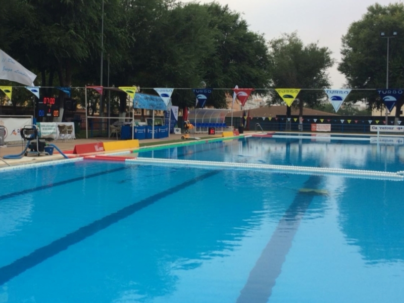 Piscina de natación con banderas y un reloj de piscina en el fondo. La piscina está rodeada por árboles y una estructura de asientos en el fondo.
