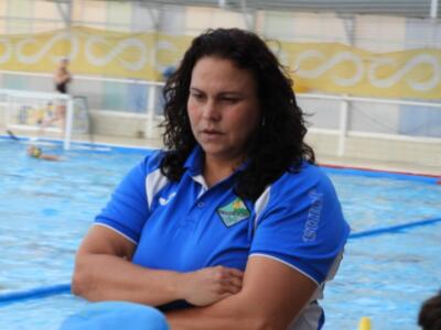 Una mujer con cabello largo y ondulado, vestida con una camiseta azul con detalles blancos, se sienta en la orilla de un estanque de natación. La piscina está rodeada por una valla blanca con un letrero amarillo y blanco. La imagen sugiere que se trata de una escena deportiva o recreativa en un complejo acuático.