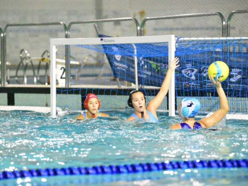 Una imagen de un partido de waterpolo femenino en una piscina. Las jugadoras están en el agua, con un portero defendiendo su portería y otras jugadores esperando para atacar. La piscina tiene líneas azules y el fondo es de color claro, probablemente blanco o gris.