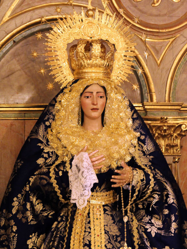 Una figura religiosa vestida con un manto negro y dorado, adornada con una corona y una guirnalda de oro. La escena se desarrolla en un entorno arquitectónico con detalles dorados y una bóveda decorada. La figura es el centro de atención, destacándose por su vestimenta y accesorios.