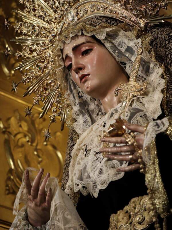 Imagen de una Virgen María vestida con trajes tradicionales religiosos, incluyendo un manto de encaje y una corona dorada con estrellas. La escena se centra en la expresión serena y digna de la figura, con un fondo dorado que resalta su presencia.