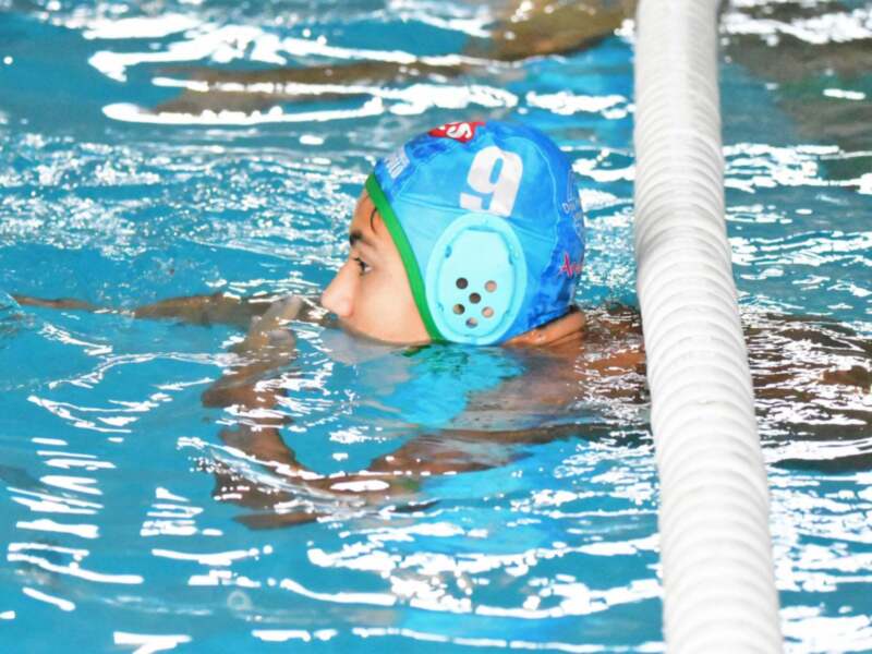 Un niño nadando en una piscina con un casco de natación azul. La imagen muestra a la persona en movimiento, con el agua ligeramente ondulada alrededor de sus pies.