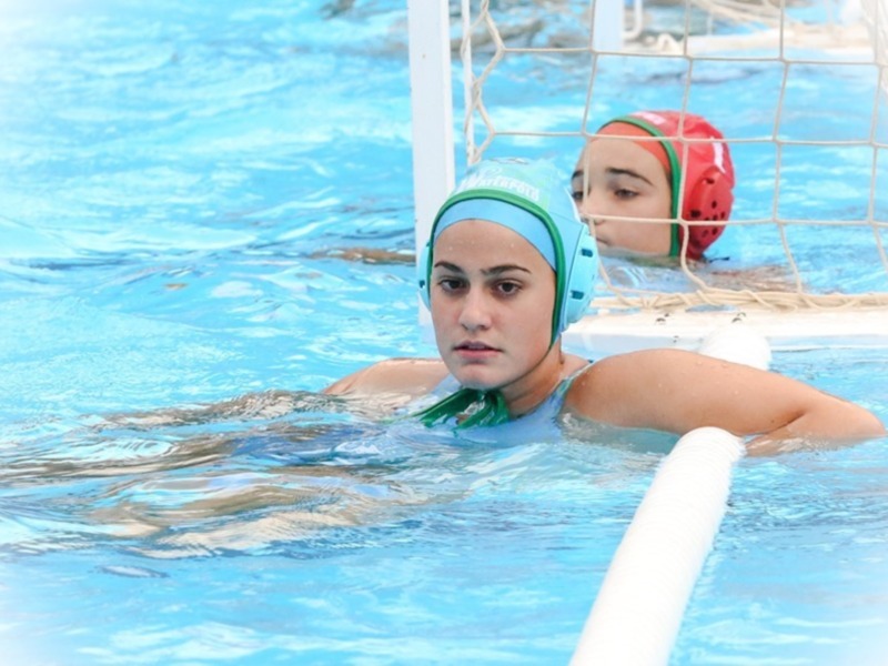 Dos jugadores de waterpolo en acción, uno con casco verde y otro con rojo, nadando en una piscina azul.