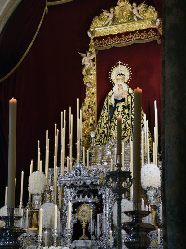 Una estatua religiosa en un altar, rodeada de velas encendidas y adornos dorados. La estatua parece ser una representación de la Virgen María, con un manto negro y detalles dorados. El fondo es oscuro con una cortina roja, enfocando la atención en el altar y la estatua.
