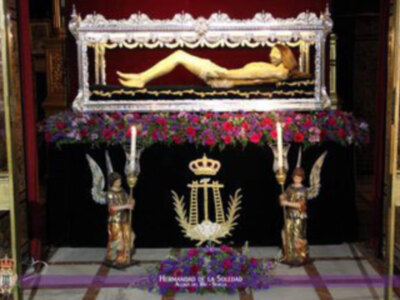 Un ataúd blanco con una figura dorada en posición de descanso, rodeado por flores rojas y blancas. En la parte superior, una corona dorada con un escudo central. En el fondo, una figura de ángel con armadura dorada y un cetro. En la parte inferior, una inscripción en letras doradas que dice "Hermandad de la Soledad".
