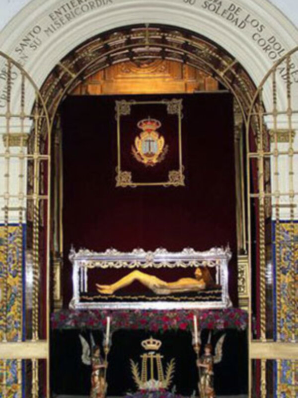 Un altar con una imagen de la Virgen María en un ataúd, rodeado por flores y decoraciones. La imagen está enmarcada con un arco de madera dorada y una corona real. La inscripción en el arco menciona "Santo Entierro de Ntra. Sra. de la Soledad".