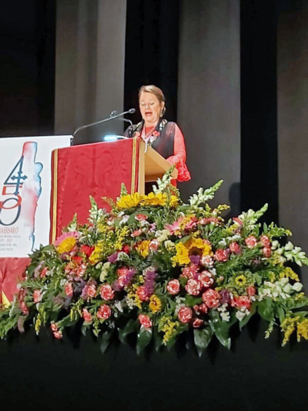 Una mujer con gafas y un vestido rojo está hablando en una mesa de conferencia. En la mesa hay flores coloridas y un cartel con el número 4. El fondo es oscuro, probablemente una sala de conferencias o auditorio.