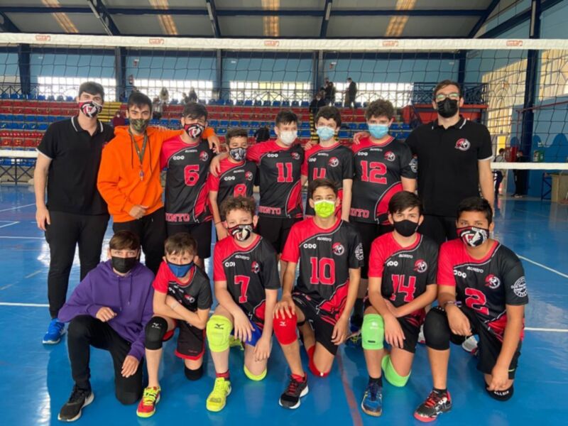 Un grupo de jóvenes jugadores de voleibol posando en un campo de juego. Todos están vestidos con camisetas rojas y negras, pantalones cortos de color rojo y calzas negras. Algunos llevan cascos protectores en los hombros, y todos están usando máscaras faciales. La imagen se toma en un gimnasio con una red de voleibol visible al fondo.