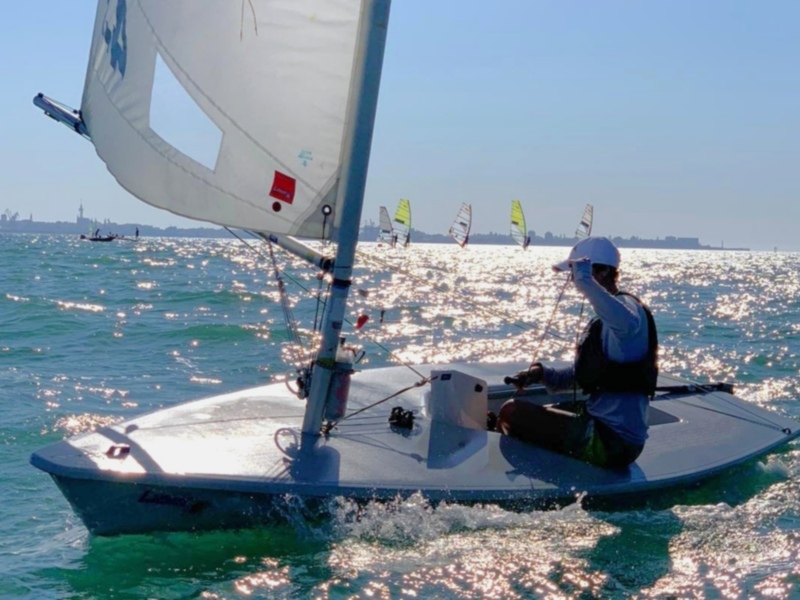 Barco de vela en el mar con un marinero al timón, banderas en el horizonte.