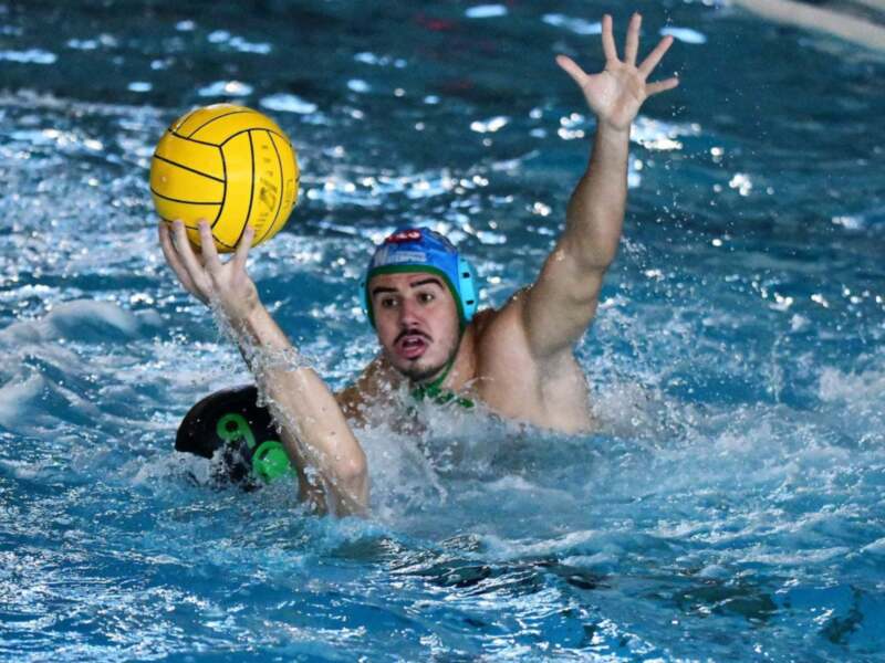 Un hombre en una piscina llena de agua azul, jugando waterpolo. Lleva un casco azul y una camiseta verde, con la mano izquierda extendida hacia un balón amarillo. La superficie del agua está llena de pequeñas burbujas y destellos, indicando la acción en el juego.