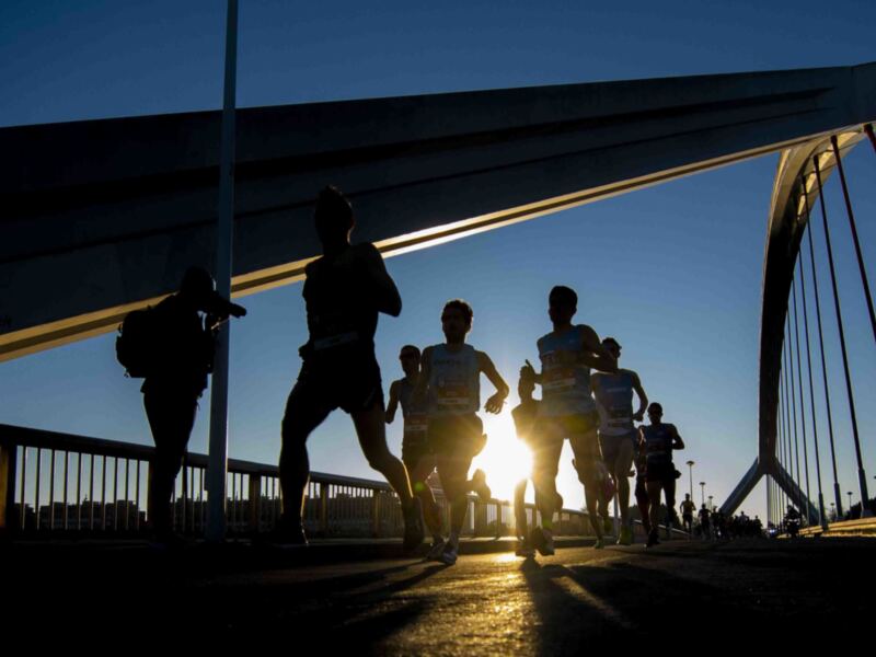 Corredores en una pista de atletismo bajo la luz del atardecer, con un puente en el fondo.