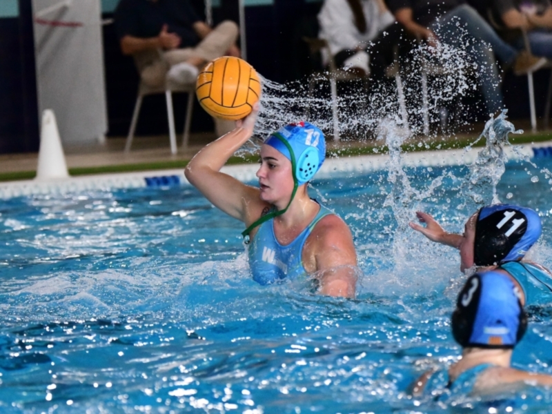 Se observa a un equipo de waterpolo en acción. Una jugadora con una gorra azul sostiene el balón mientras otra intenta alcanzarlo. La piscina es azul y transparente, con agua en movimiento, reflejando la luz del sol. La acción está intensa y dinámica, capturando el espíritu competitivo del juego.