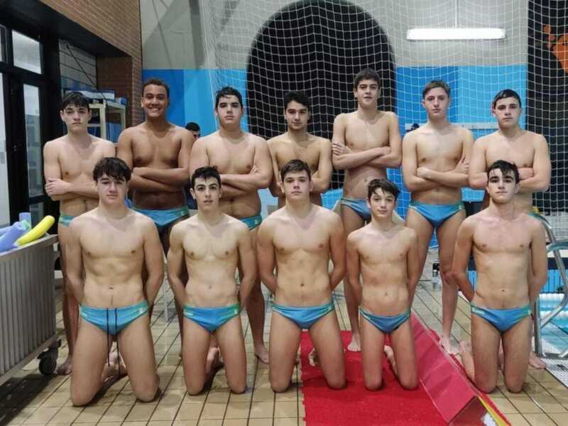 Un grupo de jóvenes vestidos con trajes de baño azul posando en una piscina. La pared de fondo es azul con cuadros blancos y hay un toldo azul detrás.