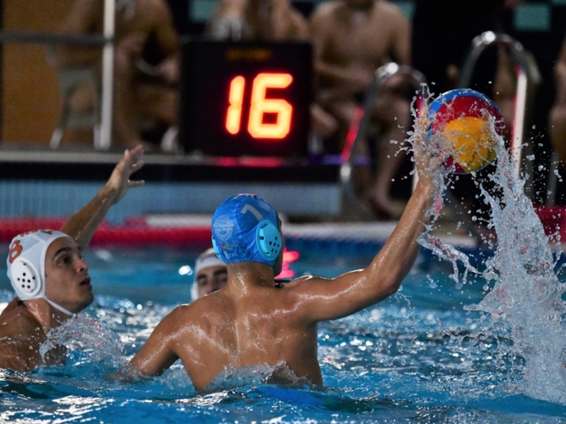 Jugadores de waterpolo en acción, con un jugador lanzando una pelota hacia el objetivo mientras otro intenta bloquearla. La imagen muestra un entorno de competición con un marcador digital que indica el tiempo restante en la final.