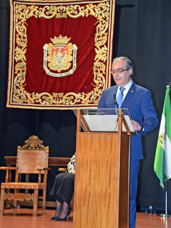 Persona hablando desde un podio en un evento formal con un escudo y silla de honor en el fondo.
