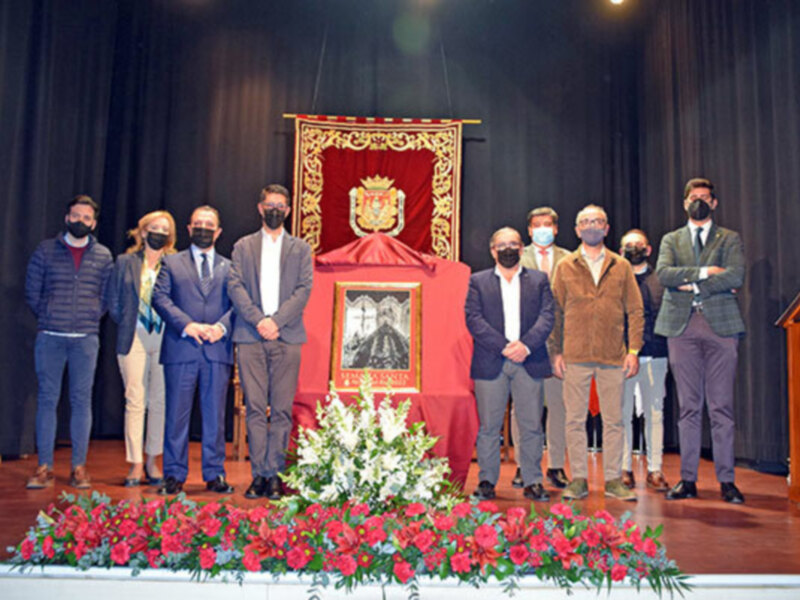 Grupo de personas con máscaras posando frente a un altar rojo con una imagen central. En el fondo, cortinas oscuras y un escudo dorado. El suelo es de madera y hay flores rojas en el frente.