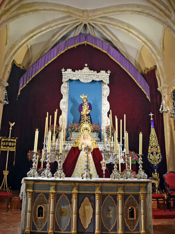 Altar con estatuas de la Virgen María y San Juan Bautista, adornado con candelabros y flores, en un interior de iglesia con techos de bóveda gótica.