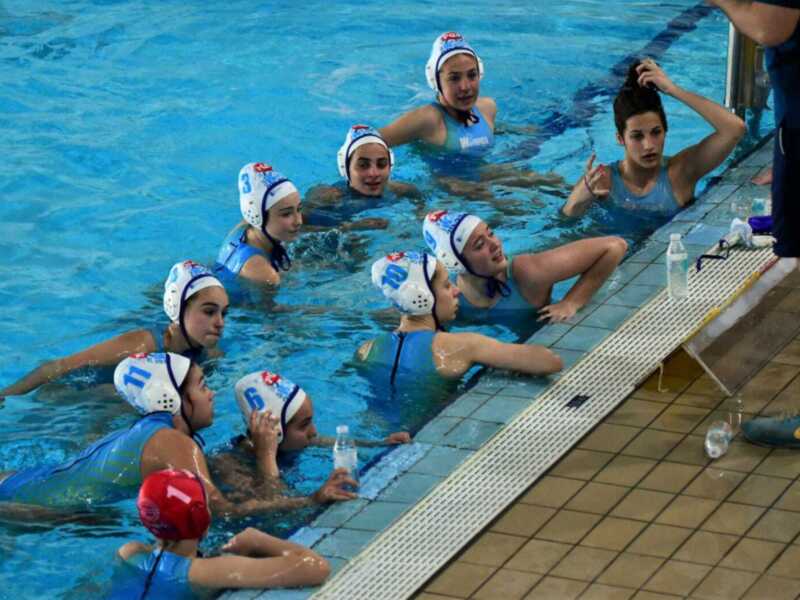 Una escena de natación en un estadio de agua con varias personas vestidas con trajes de baño y gorros de natación, posiblemente en un partido o entrenamiento.