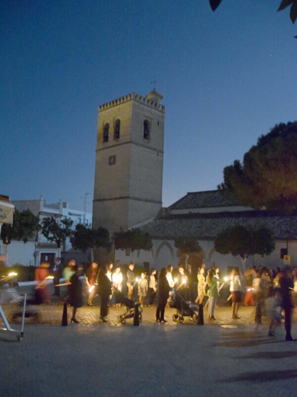 Una imagen nocturna de una plaza con un alto edificio en forma de torre, posiblemente una mezquita. La plaza está iluminada por farolas y luces de la torre, creando una atmósfera tranquila. En el fondo se pueden ver árboles y edificios más bajos, todo bajo un cielo oscuro. La imagen transmite una sensación de paz y serenidad en la noche.