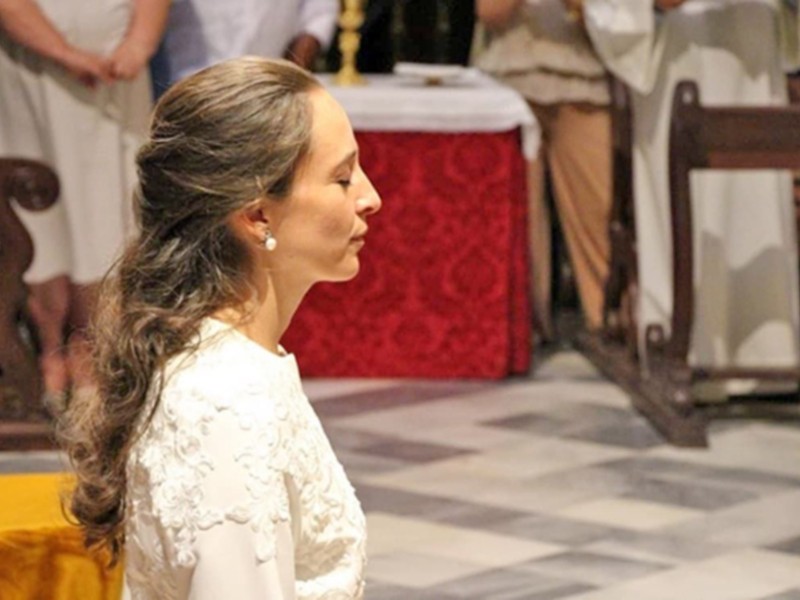 Una mujer con cabello largo y gris, vestida con un vestido blanco, se encuentra en un entorno formal con una mesa roja y un fondo con cortinas blancas y madera.
