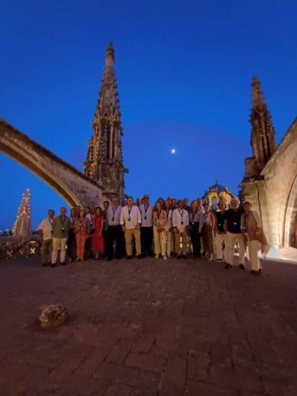Una imagen de una reunión formal en la terraza de una catedral antigua, con un grupo de personas vestidas formalmente. El cielo nocturno está claro y el sol se pone detrás de la catedral, iluminando las torres altas y ornamentadas. La escena transmite una sensación de respeto y celebración en un contexto histórico y cultural.