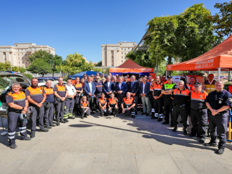 Equipo de bomberos en uniforme naranja posando juntos frente a un edificio y árboles.