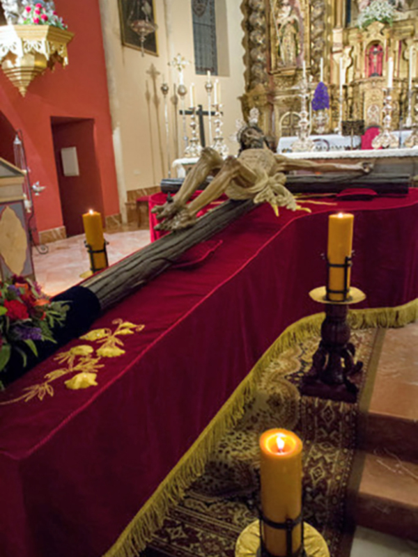 Interior de una iglesia con un altar cubierto por una manta roja y dorada. En el centro, se encuentra un crucifijo con una figura humana en posición de oración. A ambos lados, hay velas encendidas y una estatua religiosa en el fondo. La pared adyacente está pintada de rojo y hay una puerta abierta al fondo.